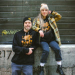 Two smiling people sporting Centraal Merch including the Mushroom Tap Hoodie and Unity Beanie