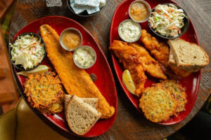 Two Buckatabon Friday fish fry entrees on red plates with coleslaw, potato pancakes, rye bread, applesauce, tartar sauce, and a lemon wedge